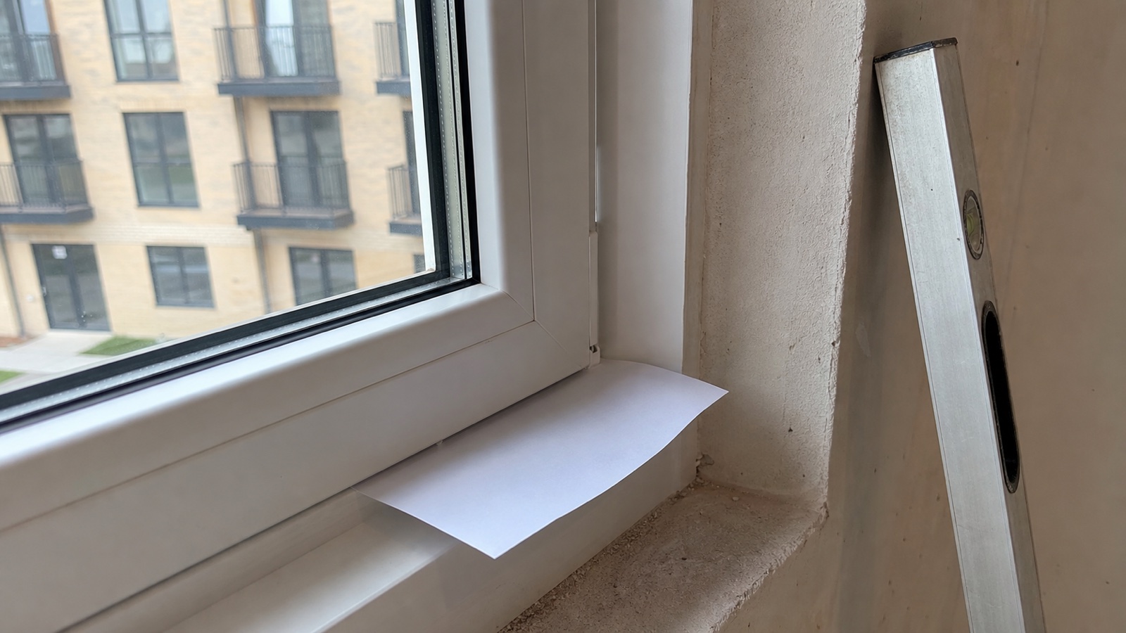 Приёмка окна в новостройке: к притвору оконной створки прижат лист бумаги для проверки на продувание, рядом уровень у откоса